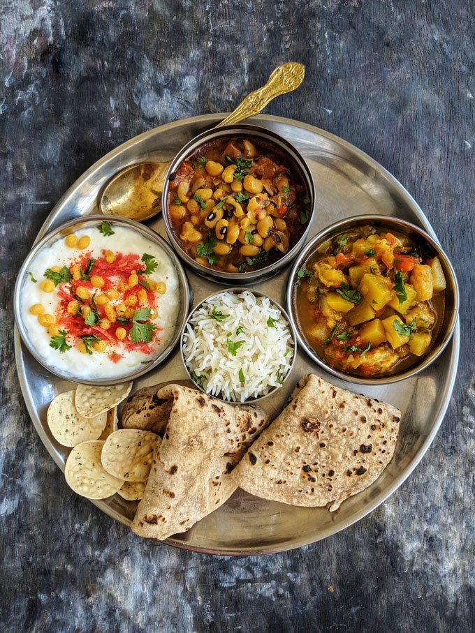 Overhead view of a simple housewarming dinner thali on a metal plate: two chapatis, a mound of steamed rice, a bowl of spiced curd garnished with chickpeas and cilantro, a bowl of black‑eyed pea curry, and a potato vegetable curry