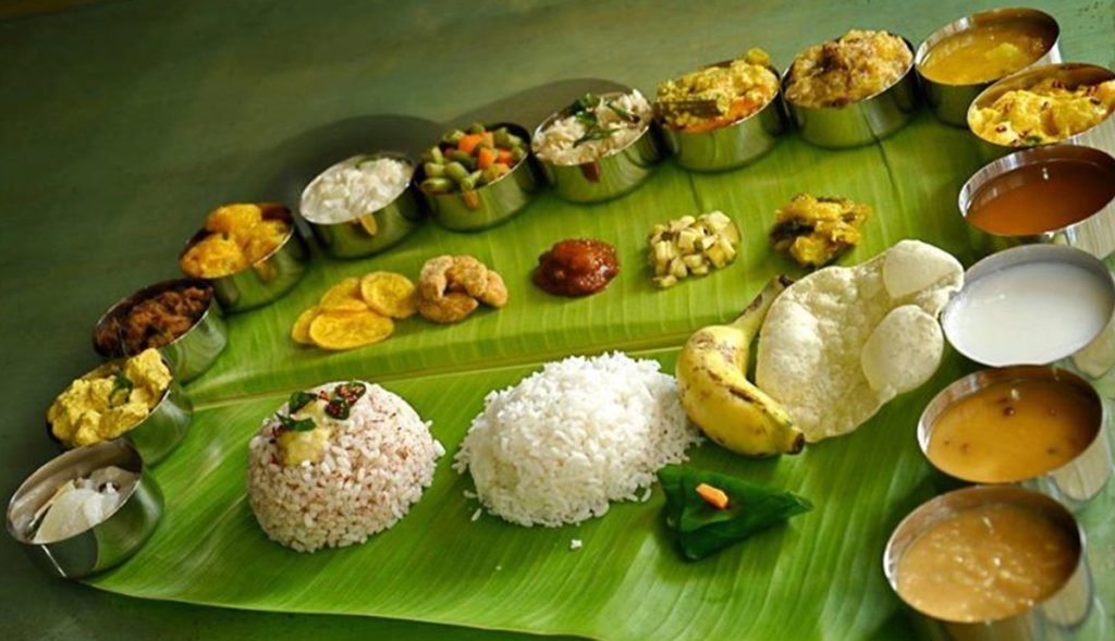 South Indian housewarming catering menu on a banana leaf featuring steamed rice, curd rice, banana, papad, and an array of small bowls with sambar, rasam, curries, vegetable sides, chutneys, and sweets